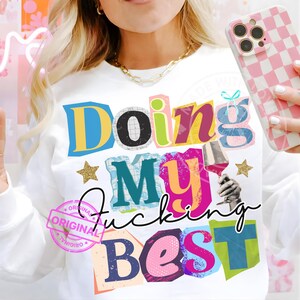 Könnte beinhalten: Weißes Sweatshirt mit dem Schriftzug "Doing My Fucking Best" in bunten Buchstaben im Collage-Stil. Das Design beinhaltet einen goldenen Stern und eine Grafik einer Hand, die ein Weinglas hält. Eine pink-weiß karierte Handyhülle ist sichtbar.