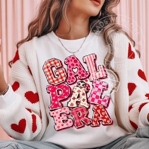 May include: A white crewneck sweatshirt featuring the text GAL PAL ERA in large, multi-patterned pink and red letters. The letters include designs like plaid, hearts, and floral prints. A small cartoon ladybug is positioned within the letter A.