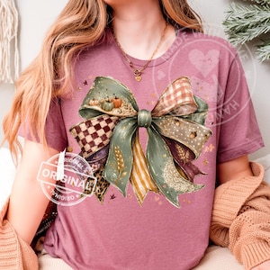Könnte beinhalten: Ein malvenfarbenes Kurzarm-T-Shirt mit einer großen zentralen Grafik einer dekorativen Schleife. Die Schleife besteht aus verschiedenen gemusterten Bändern in Salbeigrün, Braun und Creme, akzentuiert mit kleinen Kürbissen, Eicheln und Weizenähren.