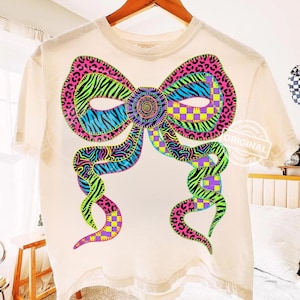 Puede incluir: Camiseta color crema con un gran diseño de lazo colorido. El lazo incorpora varios patrones, incluyendo estampado de leopardo, rayas de cebra y cuadros, en tonos vibrantes de rosa, azul, verde y morado. La camiseta está colgada en una percha de madera.