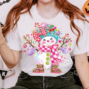 May include: A white t-shirt featuring a colorful graphic of a snowman wearing a teal hat, a pink and white checkered scarf, and leopard print boots. The snowman is decorated with red bows, candy canes, cherries, and ornaments.