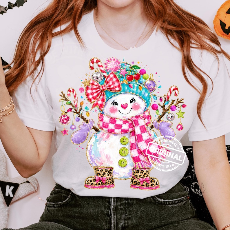 May include: A white t-shirt featuring a colorful graphic of a snowman wearing a teal hat, a pink and white checkered scarf, and leopard print boots. The snowman is decorated with red bows, candy canes, cherries, and ornaments.