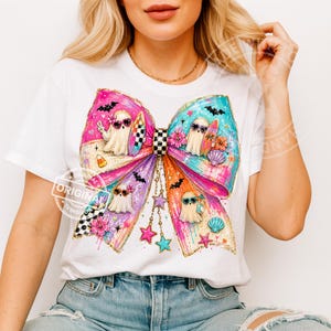 Pu&ograve; includere: T-shirt bianco con un grande design a fiocco colorato. Il fiocco presenta personaggi fantasma con occhiali da sole e tavole da surf. Il design include un centro a scacchi e accenti di stelle e conchiglie. Lo schema dei colori incorpora tonalit&agrave; rosa, blu, arancione e viola.