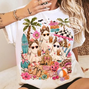 May include: White t-shirt featuring a whimsical beach scene with ghosts wearing sunglasses and holding coffee cups. The design includes a surfboard, sandcastle, pumpkins, and the text "Spooky Club" and "Beach Boo-ty".