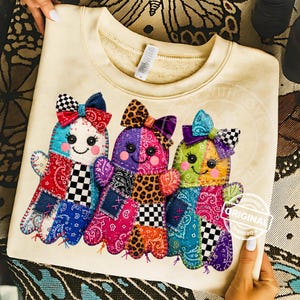 May include: A cream-colored crewneck sweatshirt featuring a graphic of three patchwork-style ghosts. Each ghost is decorated with colorful patterns including paisley, leopard print, and checkerboard, and wears a matching bow on its head.