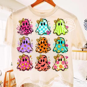 Op de afbeelding: Een crèmekleurig t-shirt met een raster van negen spoken, elk met een gouden strik. De spoken hebben diverse patronen zoals luipaardprint, ruitjes, strepen, hartjes, smileys, maïskorrels, vleermuizen en bloemen, geaccentueerd met sterren.