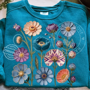 Puede incluir: Sudadera de color turquesa con diseño floral bordado. Las flores son de varios colores, incluyendo melocotón, morado y azul, con costuras detalladas. La sudadera tiene la palabra "ORIGINAL" impresa.