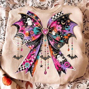 May include: A beige sweatshirt featuring a large, colorful graphic of a bow. The bow is decorated with ghosts, bats, stars, moons, flowers, and a central pink heart. Dangling chains with charms, including bats and crystals, hang from the bow.