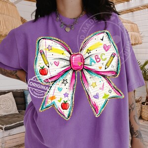 May include: A purple t-shirt featuring a large, colorful bow design. The bow is decorated with school-themed elements like pencils, apples, hearts, stars, and the letters "ABC". The design has a watercolor effect with gold glitter accents.