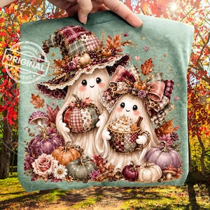 May include: A sage green t-shirt featuring a graphic of two white ghosts. One ghost wears a plaid witch hat, the other a large plaid bow. They are surrounded by colorful pumpkins, autumn leaves, mushrooms, and a mug of hot cocoa with whipped cream.