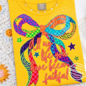 Puede incluir: Camiseta amarilla con un diseño de lazo colorido y el texto "All my life you have been faithful." El lazo presenta varios patrones, incluyendo estampado de leopardo y rayas de cebra. Se ve un collar pequeño en la parte superior.