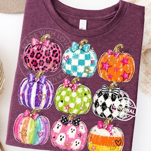 May include: A maroon short-sleeved t-shirt featuring a graphic print of eight stylized pumpkins. The pumpkins display various patterns including leopard print, checkered, polka dots, and stripes, each topped with a decorative bow and small accents.