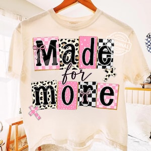 May include: Cream-colored t-shirt with the phrase "Made for More" in a playful, colorful design. The text is arranged in blocks with pink, black and white patterns, and a cross graphic. The shirt is hanging on a wooden hanger.