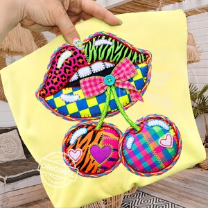 Puede incluir: Un cojín amarillo con un diseño de aplique colorido que presenta unos labios y dos cerezas. Los labios tienen un estampado de leopardo rosa, rayas de cebra y un patrón de cuadros azul y amarillo. Las cerezas tienen corazones y un patrón de cuadros.