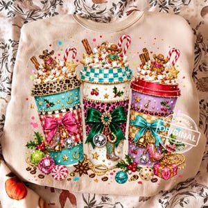 May include: A beige sweatshirt featuring a graphic of three coffee cups topped with whipped cream, marshmallows, candy canes, and gingerbread figures. The cups are decorated with bows, leopard print, stars, and western-themed charms.