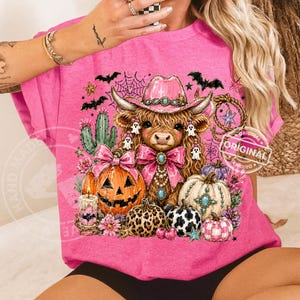 Puede incluir: Una camiseta rosa con un dise&ntilde;o de Halloween. El gr&aacute;fico presenta una vaca de las Tierras Altas con un sombrero de vaquero rosa, rodeada de calabazas, murci&eacute;lagos y fantasmas. El dise&ntilde;o tiene una est&eacute;tica festiva y caprichosa.