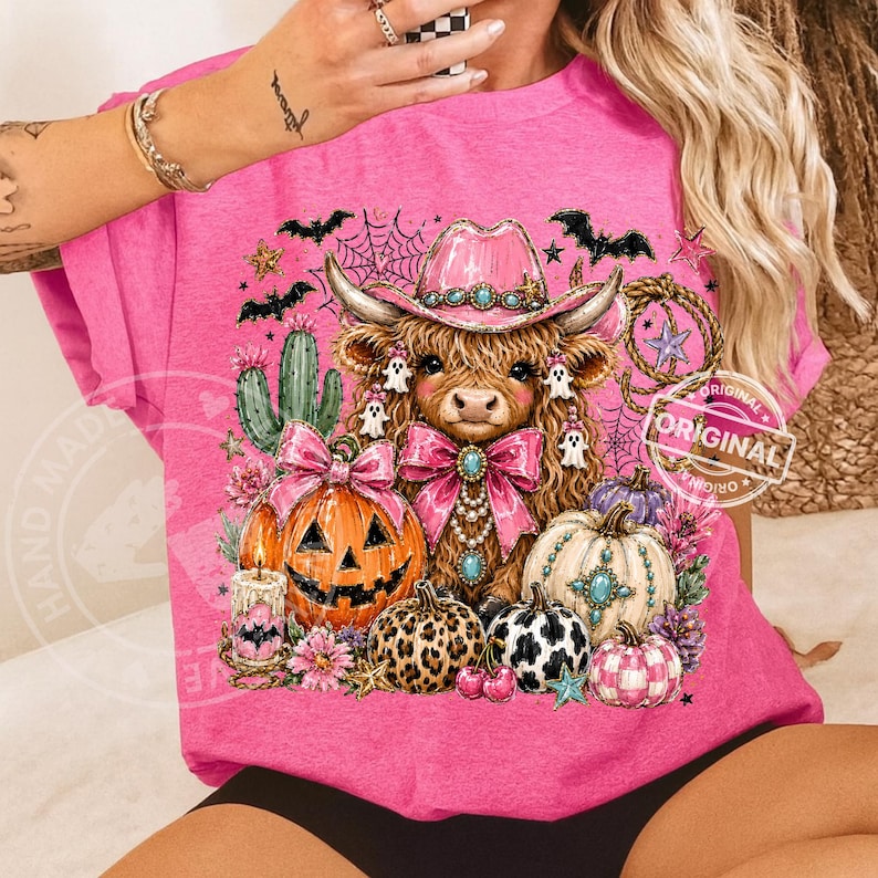 Puede incluir: Una camiseta rosa con un dise&ntilde;o de Halloween. El gr&aacute;fico presenta una vaca de las Tierras Altas con un sombrero de vaquero rosa, rodeada de calabazas, murci&eacute;lagos y fantasmas. El dise&ntilde;o tiene una est&eacute;tica festiva y caprichosa.