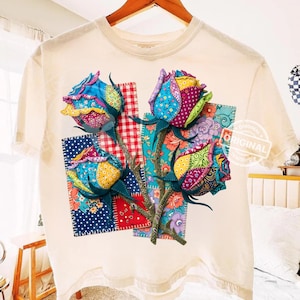 Może przedstawiać: Kremowy t-shirt z kolorowym kwiatowym wzorem. Projekt przedstawia trzy róże z płatkami w stylu patchwork w różnych kolorach, w tym niebieskim, czerwonym i żółtym. Róże umieszczone są na tle wzorzystych kwadratów.