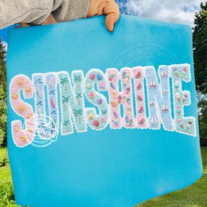 Op de afbeelding: Een turquoise blauwe strandlaken met het woord "SUNSHINE" geborduurd in een kleurrijk strandthema. De letters zijn gevuld met afbeeldingen van schelpen, surfplanken en ijsjes. De handdoek wordt omhooggehouden tegen een blauwe lucht en groen gras.