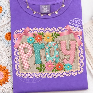 Peut inclure: Un t-shirt violet Comfort Colors avec le mot "PRAY" dans un motif floral. Les lettres sont roses, turquoise et à carreaux, entourées de fleurs et d'une bordure en dentelle. Un collier doré et un autocollant "ORIGINAL" sont également visibles.
