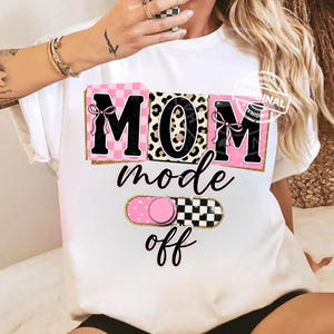 Puede incluir: Camiseta blanca con un diseño gráfico. El diseño presenta la palabra "MOM" en letras grandes, con patrones de cuadros rosas y leopardo. Debajo, las palabras "mode off" están escritas en cursiva, con un interruptor basculante rosa y negro.