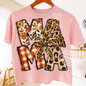 Puede incluir: Una camiseta de manga corta de color rosa claro con la palabra MAMA en letras grandes de bloque. Las letras tienen diferentes patrones: floral, estampado de leopardo y cuadros naranjas. Un lazo de leopardo est&aacute; centrado sobre la palabra.