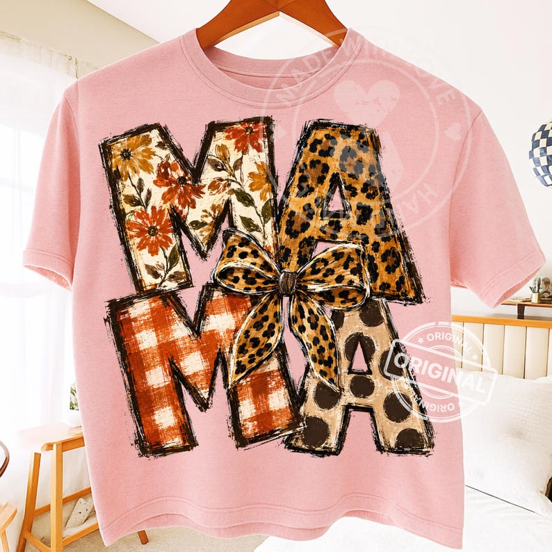 Puede incluir: Una camiseta de manga corta de color rosa claro con la palabra MAMA en letras grandes de bloque. Las letras tienen diferentes patrones: floral, estampado de leopardo y cuadros naranjas. Un lazo de leopardo est&aacute; centrado sobre la palabra.