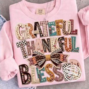 Könnte beinhalten: Ein hellrosa Sweatshirt mit Rundhalsausschnitt und den Wörtern GRATEFUL THANKFUL BLESSED in großen, gemusterten Buchstaben. Das Design enthält eine zentrale Schleife mit Leopardenmuster und Karomustern, umgeben von Blumen- und Geometriemotiven.