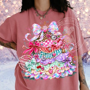 Puede incluir: Una camiseta de color rosa empolvado con un gran gráfico colorido de un árbol de Navidad estilizado compuesto por volantes superpuestos, lazos, estampado de leopardo, cuadros, bolas de discoteca, cerezas y adornos en forma de corazón.