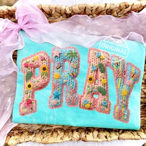May include: A turquoise t-shirt featuring the word "PRAY" embroidered with a floral design. The letters are outlined in pink and decorated with sunflowers, butterflies, and other embellishments. The shirt is placed in a woven basket.