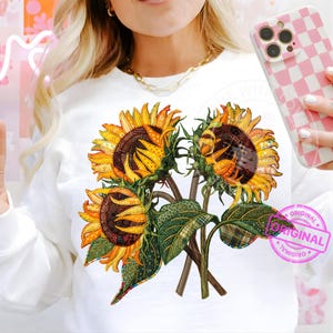 May include: White sweatshirt featuring a vibrant sunflower bouquet design. The sunflowers have yellow petals and brown centers, with green leaves and stems. The design includes plaid and floral patterns. A pink stamp with the word "ORIGINAL" is visible.