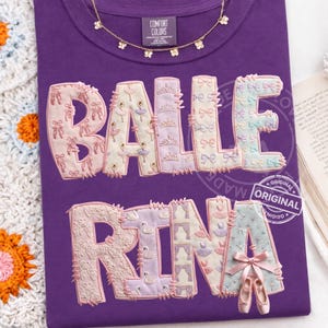 May include: A purple t-shirt with the words "BALLERINA" in a decorative, embroidered style. The letters are filled with ballet-themed images like pointe shoes, tutus, and swans. A gold necklace with flower charms is at the neckline.