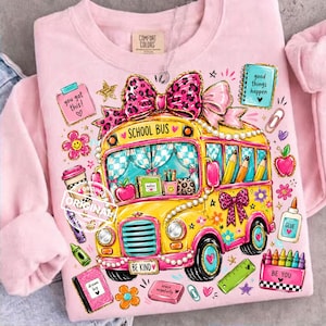 Puede incluir: Sudadera rosa con un diseño de autobús escolar amarillo, adornada con un lazo rosa y detalles de estampado de leopardo. El autobús tiene las palabras "SCHOOL BUS" y "BE KIND". Gráficos adicionales: manzanas, lápices y el texto "good things happen".