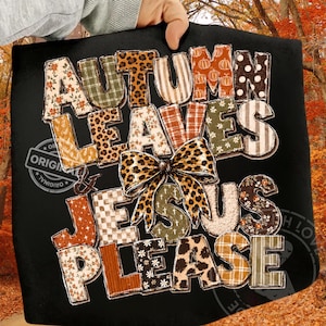 May include: A black t-shirt featuring the text AUTUMN LEAVES & JESUS PLEASE in a multi-patterned block font. The letters incorporate various prints including leopard, plaid, floral, and pumpkin motifs, centered with a large leopard print bow.