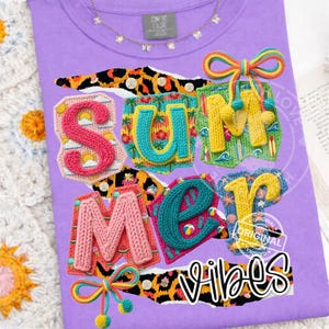 May include: A lilac Comfort Colors t-shirt with the words "SUMMER VIBES" in colorful, textured letters. The letters are embellished with beach-themed designs and a leopard print background. A gold necklace with small charms is visible.