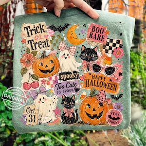 May include: A teal Halloween-themed tote bag featuring a collage of spooky illustrations. The design includes pumpkins, ghosts, black cats, and the phrases "Trick or Treat," "Happy Halloween," and "Boo Babe." The bag is held by a hand.