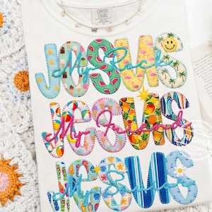 May include: White Comfort Colors t-shirt with the words "Jesus My Rock, Jesus My Friend, Jesus Saves" in colorful, patterned letters. The letters feature designs like watermelon, ice cream, and flamingos. A necklace is visible at the neckline.
