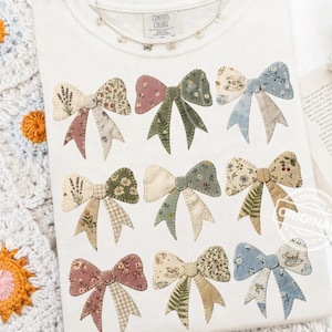Op de afbeelding: Wit t-shirt met negen stoffen strikapplicaties in verschillende bloemen- en patroonontwerpen. De strikken zijn in tinten roze, groen, blauw en beige. Het shirt heeft de tekst "COMFORT COLORS" op het label.