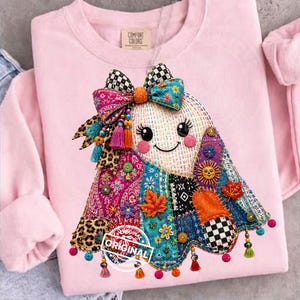 May include: A light pink sweatshirt featuring a central graphic of a ghost adorned with a large checkered bow, colourful tassels, and a patchwork-style pattern including leopard print, floral motifs, maple leaves, and a sun, with the word ORIGINAL.