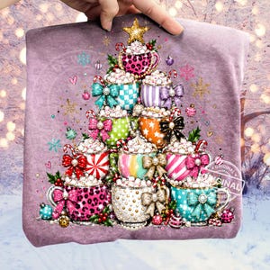 Pode incluir: Uma t-shirt malva com uma impressão gráfica de uma árvore de Natal construída a partir de canecas empilhadas cheias de chantilly. As canecas exibem vários padrões, incluindo leopardo, riscas e bolinhas, acentuadas com laços e enfeites.
