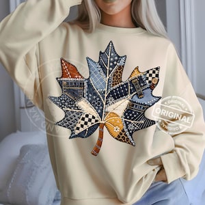 May include: A cream-colored long-sleeve sweatshirt featuring a large central graphic of a maple leaf. The leaf design is composed of various patchwork patterns including denim textures, leopard print, checkered squares, and floral motifs.