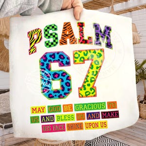 May include: A white blanket with the text "PSALM 67" in colorful, patterned letters. Below, the text reads "MAY GOD BE GRACIOUS TO US AND BLESS US AND MAKE HIS FACE SHINE UPON US."
