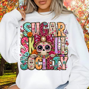 Może przedstawiać: Biała bluza z długim rękawem z kolorowym nadrukiem graficznym z napisem "SUGAR SKULL SOCIETY". Wzór przedstawia centralnie umieszczoną czaszkę cukrową z dużą różową kokardą w panterkę, otoczoną motywami kwiatowymi, motylami i sercami.