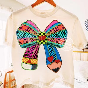 May include: Cream-coloured t-shirt with a large, colourful bow design. The bow incorporates zebra stripes, leopard print, and hearts in vibrant pink, blue, and green. A pencil design is at the bottom of the bow.