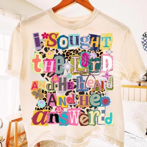 Può includere: T-shirt color crema con un design a collage colorato. Il design presenta il testo "I Sought The Lord And He Heard And He Answered" in vari font e colori, con accenti floreali e faccine sorridenti.