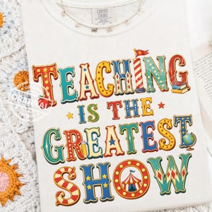 Könnte beinhalten: Cremefarbenes T-Shirt mit dem Text "TEACHING IS THE GREATEST SHOW" in farbenfrohen Buchstaben im Zirkusstil. Das Design umfasst Sterne, einen Globus, einen Bleistift und ein Zirkuszelt. Das Shirt hat ein Comfort Colors-Etikett.