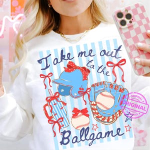 May include: White sweatshirt with a baseball theme. The design includes a blue baseball helmet, glove, and baseball, with red bows and the text "Take me out to the Ballgame" in blue script. The background has blue and white stripes.