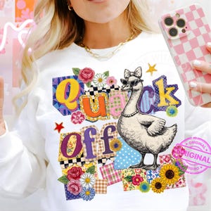 May include: White sweatshirt with the phrase "Quack Off" in colorful patchwork letters and a drawing of a goose wearing sunglasses and a pearl necklace. Floral and star accents surround the text and goose.