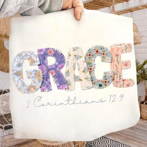 May include: A white canvas featuring the word "GRACE" in large, colourful, patchwork-style letters. Each letter is decorated with floral patterns in shades of purple, blue, and pink. The text "2 Corinthians 12:9" is written below.