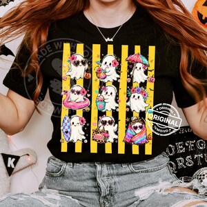 May include: A black t-shirt featuring a graphic print of nine white ghosts in various summer-themed activities, arranged over vertical yellow stripes. The ghosts are depicted with accessories like sunglasses, floral crowns, a surfboard, and a beach bag.